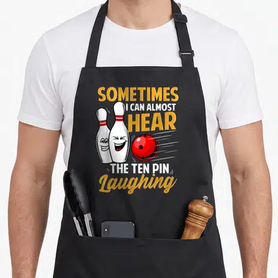 Almost pins for aprons - Two laughing cartoon bowling pins and red ball with yellow and white text — shown on a black cotton canvas apron against a white background