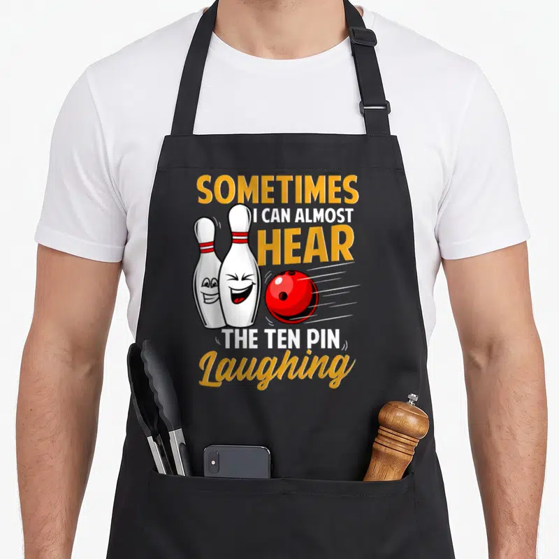 Almost pins for aprons - Two laughing cartoon bowling pins and red ball with yellow and white text — shown on a black cotton canvas apron against a white background