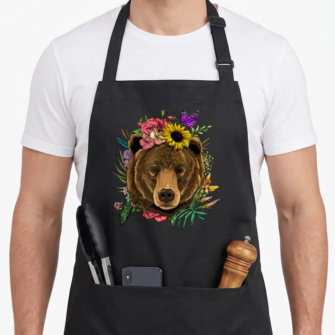 Bear floral apron - Brown bear head surrounded by yellow sunflowers, pink roses, and a purple butterfly perched on top — shown on a black cotton canvas apron against a white background