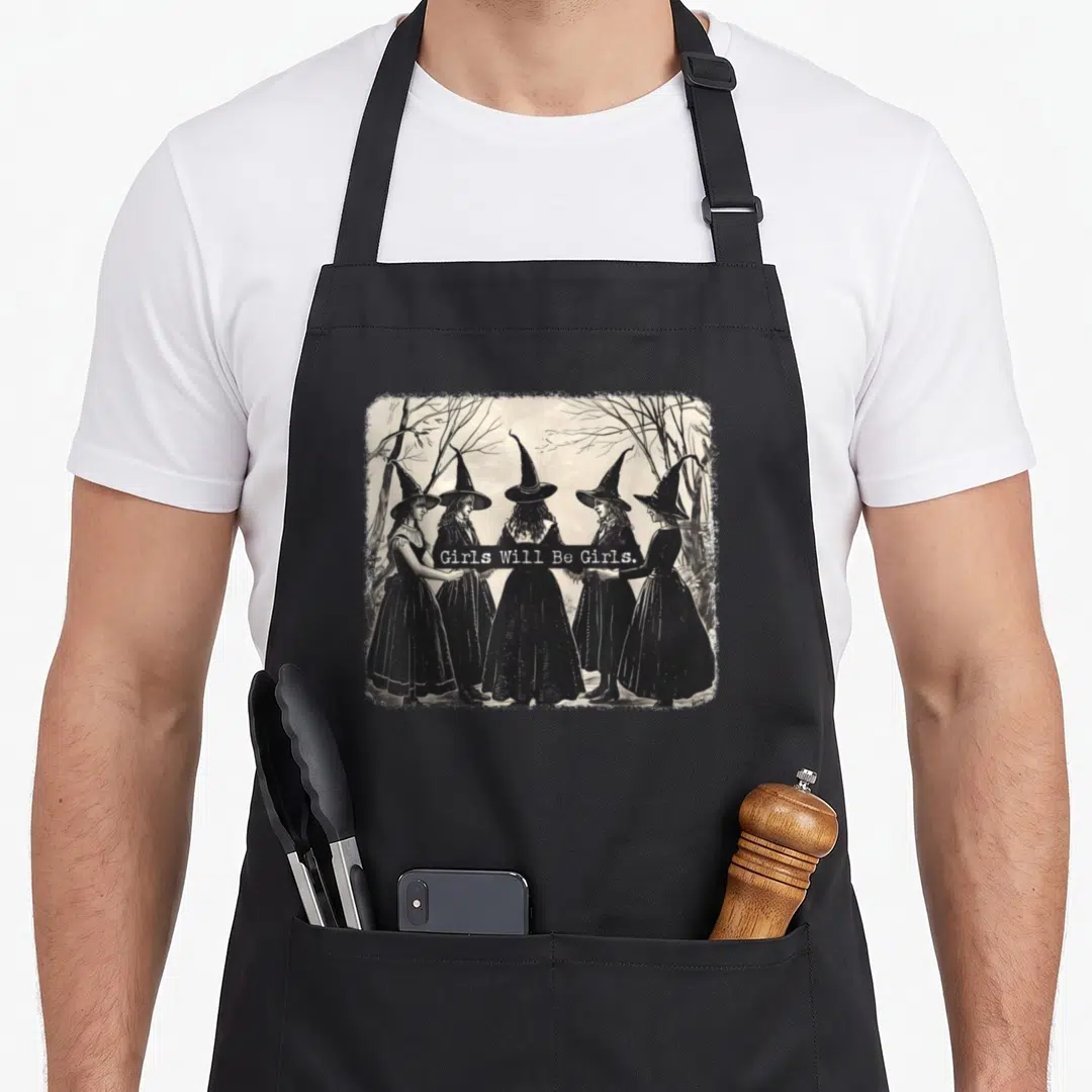 Witchy witch apron - Five witches in black dresses holding a white banner reading Girls Will Be Girls in a grey wooded — shown on a black cotton canvas apron against a white background