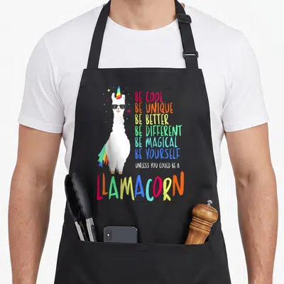 Llamacorn unicorn apron - Rainbow horned llama wearing sunglasses next to stacked colorful text reading Be Cool Be — shown on a black cotton canvas apron against a white background