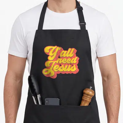 Jesus 70s christian apron - Yellow cursive text reading Y all need Jesus with a red shadow background — shown on a black cotton canvas apron against a white background