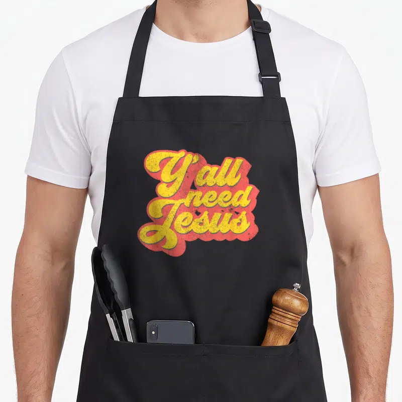 Jesus 70s christian apron - Yellow cursive text reading Y all need Jesus with a red shadow background — shown on a black cotton canvas apron against a white background