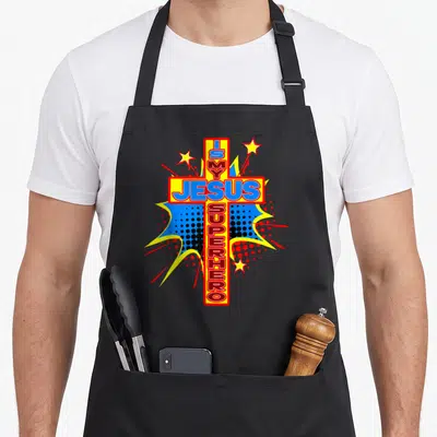 Church superhero apron - Red cross with J IS MY JESUS SUPERHERO text and three stars in comic style — shown on a black cotton canvas apron against a white background