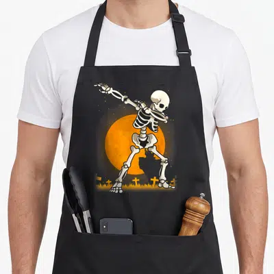 Dabbing skeleton halloween apron - Cartoon skeleton dabbing pose in front of orange moon with small crosses and grass — shown on a black cotton canvas apron against a white background