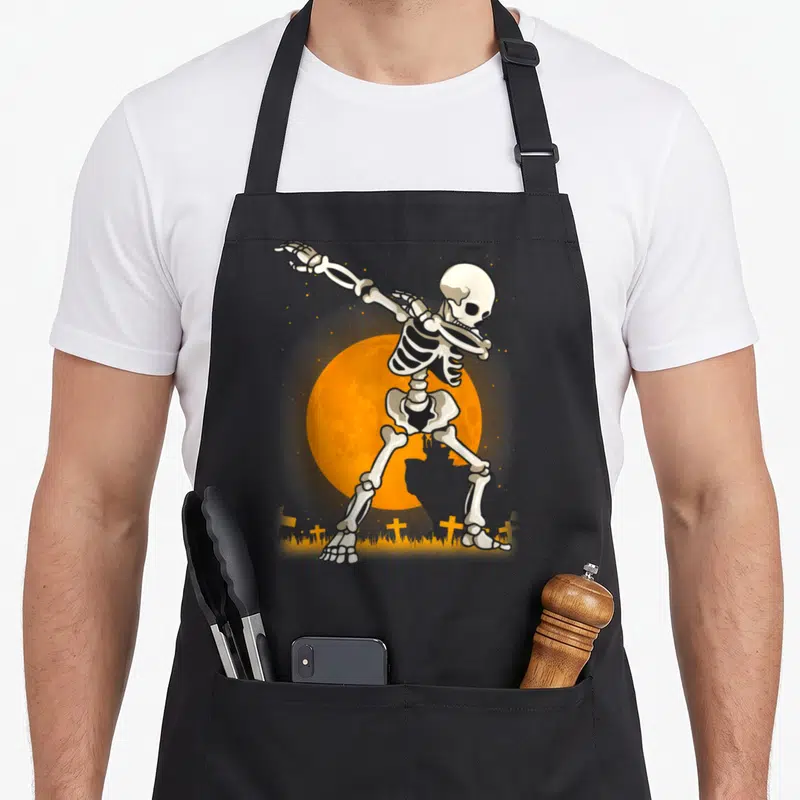 Dabbing skeleton halloween apron - Cartoon skeleton dabbing pose in front of orange moon with small crosses and grass — shown on a black cotton canvas apron against a white background