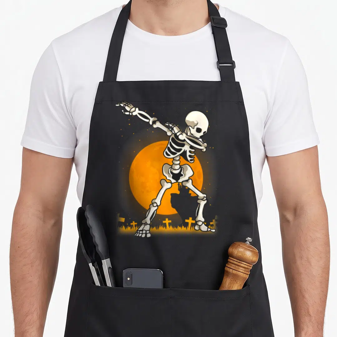Dabbing skeleton halloween apron - Cartoon skeleton dabbing pose in front of orange moon with small crosses and grass — shown on a black cotton canvas apron against a white background