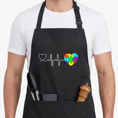 Acceptance autism awareness apron - White heartbeat line connects outline heart to multi-colored puzzle piece heart — shown on a black cotton canvas apron against a white background