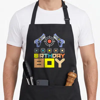 Laser birthday apron - Two blue laser guns flanking a red target above five small targets and BIRTHDAY BOY text — shown on a black cotton canvas apron against a white background