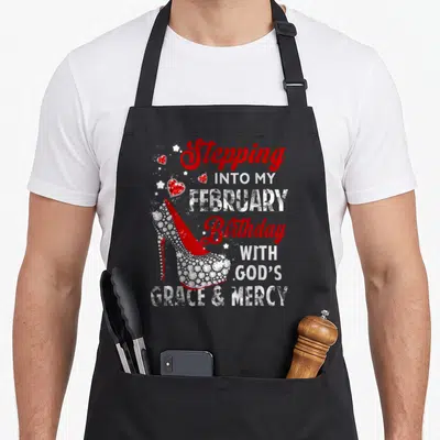 Stepping february birthday apron - Red high heel shoe with stacked Stepping into My February Birthday With Gods Grace — shown on a black cotton canvas apron against a white background
