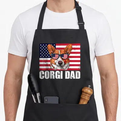 Flag corgi aprons - Distressed corgi head with flag sunglasses over USA flag, 'Corgi Dad' text below in white — shown on a black cotton canvas apron against a white background
