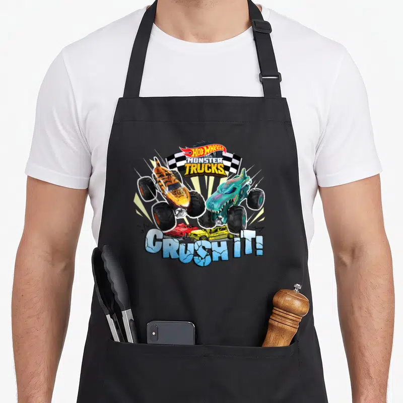 Monster wheel apron - Two monster trucks jumping over crushed cars with text CRUSH IT and checkered flag — shown on a black cotton canvas apron against a white background