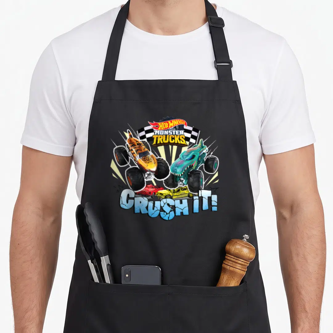 Monster wheel apron - Two monster trucks jumping over crushed cars with text CRUSH IT and checkered flag — shown on a black cotton canvas apron against a white background