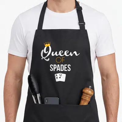 Game queen apron - Queen OF SPADES text with crown and two playing cards Ace and Queen of Spades — shown on a black cotton canvas apron against a white background