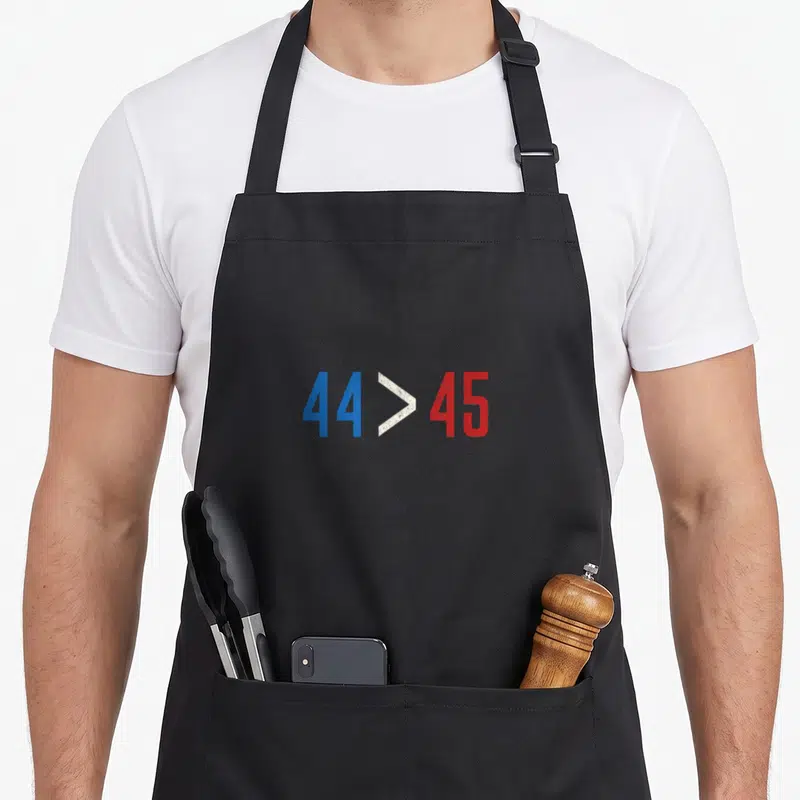 Greater midterms apron - Distressed blue 44 and red 45 numbers with white greater than symbol — shown on a black cotton canvas apron against a white background