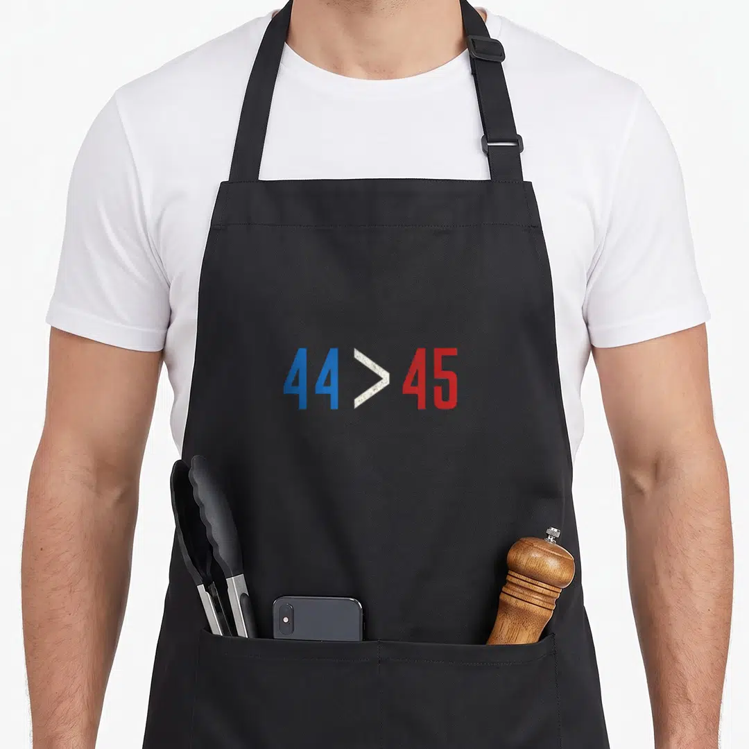 Greater midterms apron - Distressed blue 44 and red 45 numbers with white greater than symbol — shown on a black cotton canvas apron against a white background
