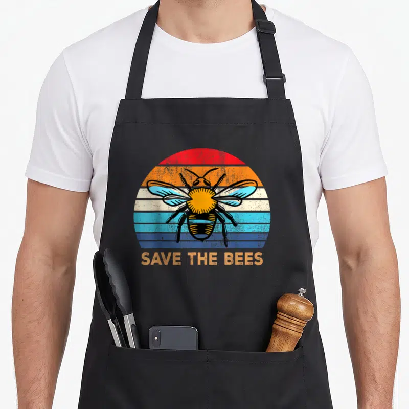 Save bee change apron - Retro bee illustration on striped semi-circle with SAVE THE BEES text below — shown on a black cotton canvas apron against a white background