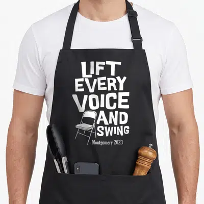 Montgomery folding aprons - White stacked text Lift Every Voice And Swing with line-art folding chair and Montgomery — shown on a black cotton canvas apron against a white background