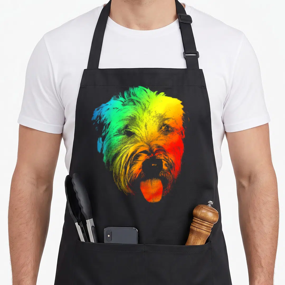 Coated dog dad apron - Rainbow gradient portrait of a wheaten terrier head with tongue out in blue, green, and red — shown on a black cotton canvas apron against a white background