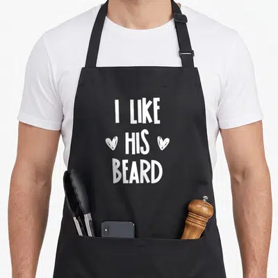 Congratulation beard apron - White text I LIKE HIS BEARD with two hearts flanking the middle word on black fabric — shown on a black cotton canvas apron against a white background