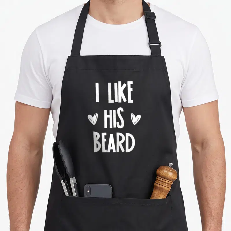 Congratulation beard apron - White text I LIKE HIS BEARD with two hearts flanking the middle word on black fabric — shown on a black cotton canvas apron against a white background