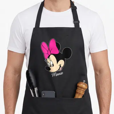 Vintage face friends apron - Cartoon Minnie Mouse head with pink bow and beige face, text Minnie below — shown on a black cotton canvas apron against a white background