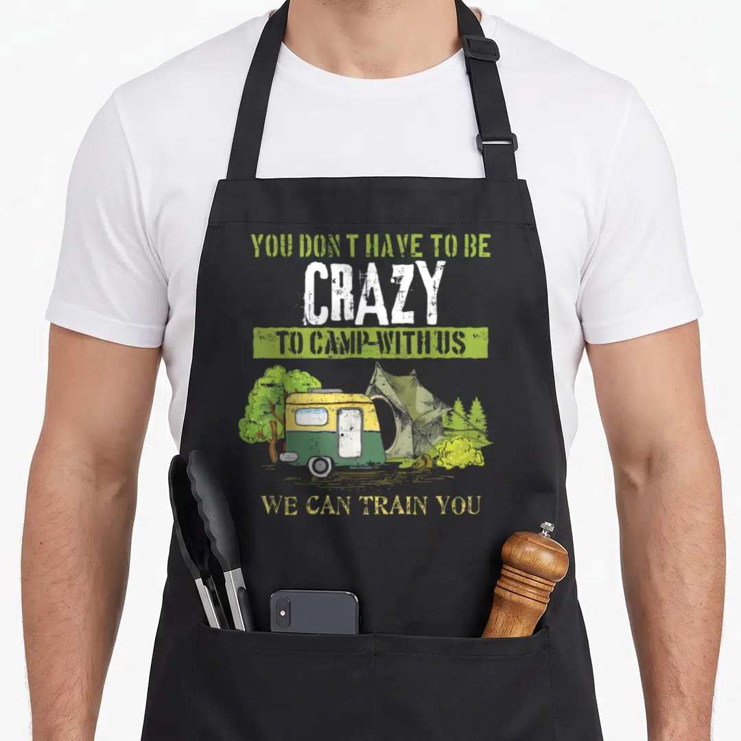 Joke camping apron - Distressed camper and tent illustration with You Don't Have To Be Crazy To Camp With Us We Can — shown on a black cotton canvas apron against a white background