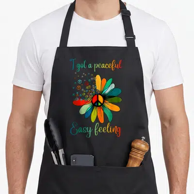 Feeling sunflower apron - Multi-colored flower with peace symbols and text reading I got a peaceful Easy feeling — shown on a black cotton canvas apron against a white background