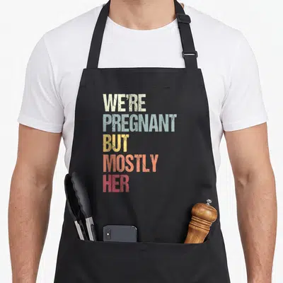 Pregnant dad but apron - Stacked text reading We're Pregnant But Mostly Her in beige, blue, yellow, coral, and red — shown on a black cotton canvas apron against a white background