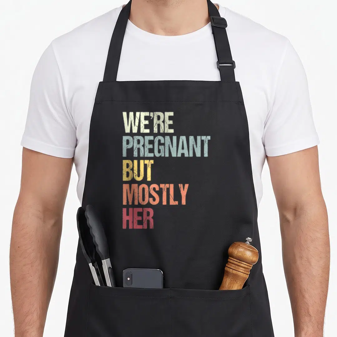Pregnant dad but apron - Stacked text reading We're Pregnant But Mostly Her in beige, blue, yellow, coral, and red — shown on a black cotton canvas apron against a white background
