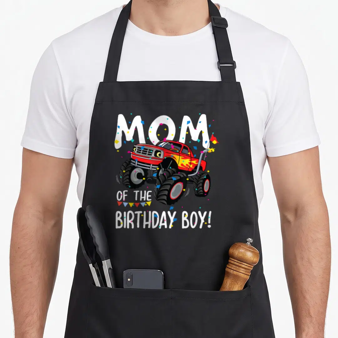 Monster birthday truck apron - Red monster truck with MOM OF THE BIRTHDAY BOY text and confetti in white red blue yellow — shown on a black cotton canvas apron against a white background