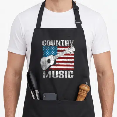 Flag guitarist cook's country aprons - Distressed COUNTRY MUSIC text framing a white guitar over a red white and blue — shown on a black cotton canvas apron against a white background