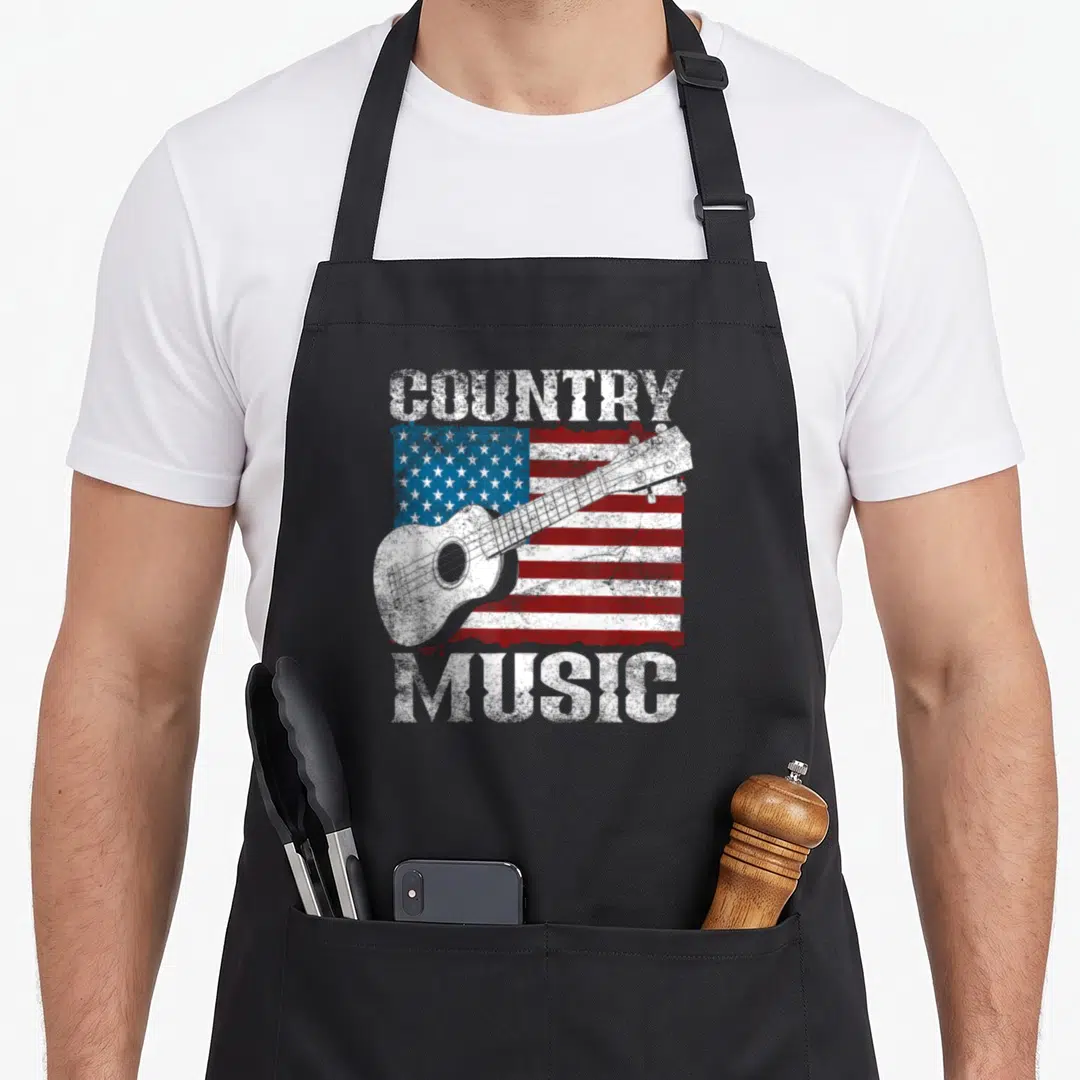 Flag guitarist cook's country aprons - Distressed COUNTRY MUSIC text framing a white guitar over a red white and blue — shown on a black cotton canvas apron against a white background