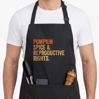 Right spice apron - Stacked PUMPKIN SPICE & REPRODUCTIVE RIGHTS text in orange peach and yellow gradients — shown on a black cotton canvas apron against a white background