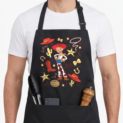 Jessie western apron - Cartoon cowgirl Jessie with SHERIFF stars, red hat, yellow cactus, and blue jeans — shown on a black cotton canvas apron against a white background