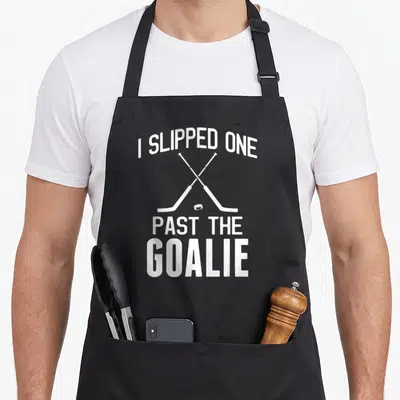 Hockey daddy apron - White I SLIPPED ONE PAST THE GOALIE text with crossed hockey sticks and puck — shown on a black cotton canvas apron against a white background