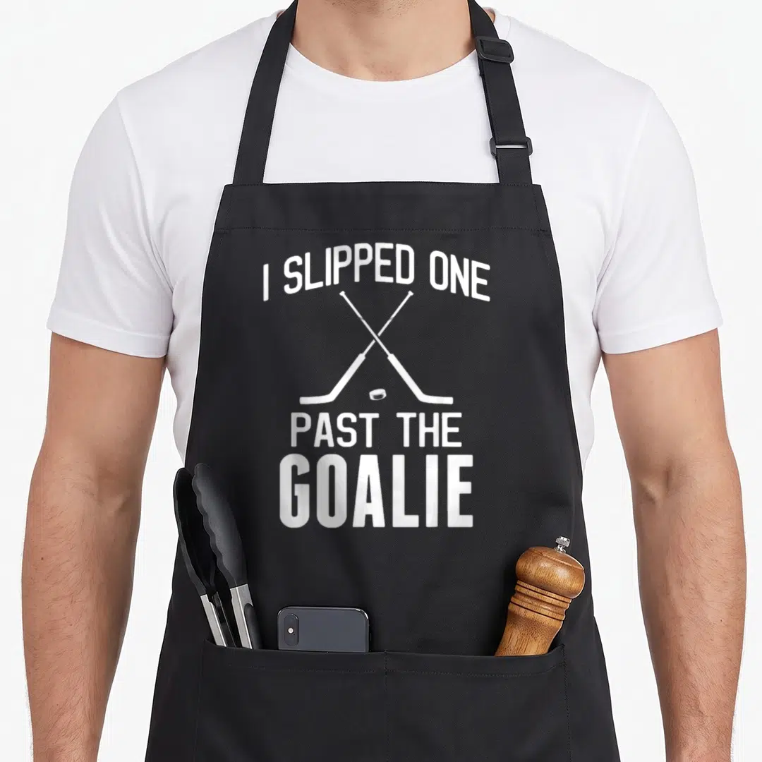 Hockey daddy apron - White I SLIPPED ONE PAST THE GOALIE text with crossed hockey sticks and puck — shown on a black cotton canvas apron against a white background