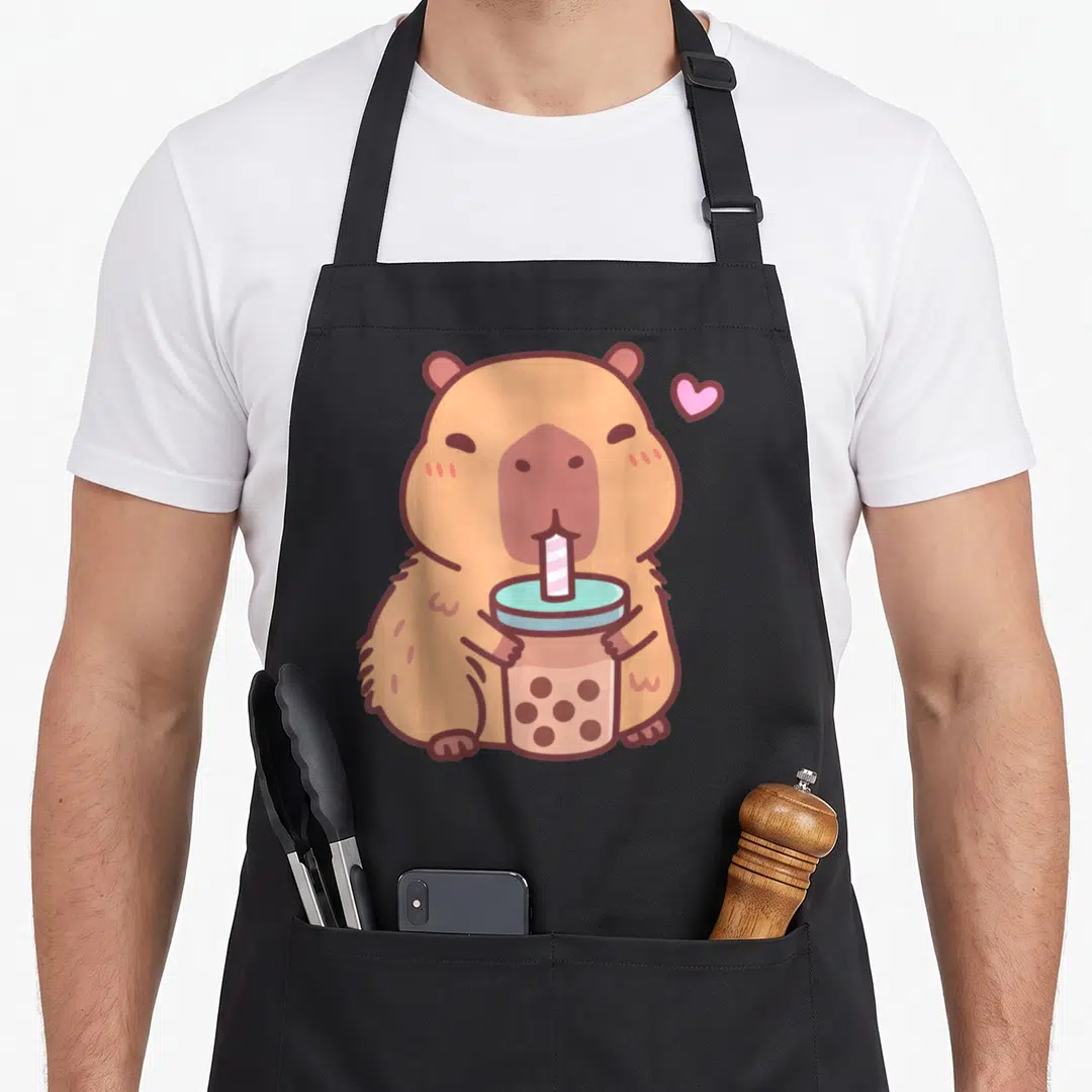 Capybara anime apron - Tan cartoon capybara holding bubble tea cup with pink heart floating nearby — shown on a black cotton canvas apron against a white background