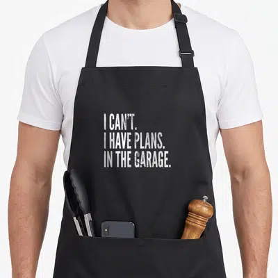 Car garage apron - White stacked text reading I CAN'T I HAVE PLANS IN THE GARAGE — shown on a black cotton canvas apron against a white background