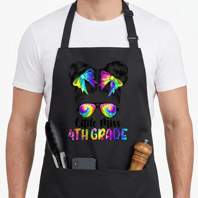 Miss 4th tie-dye apron - Cartoon girl with two buns, rainbow bows, sunglasses, and Little Miss 4TH GRADE text — shown on a black cotton canvas apron against a white background