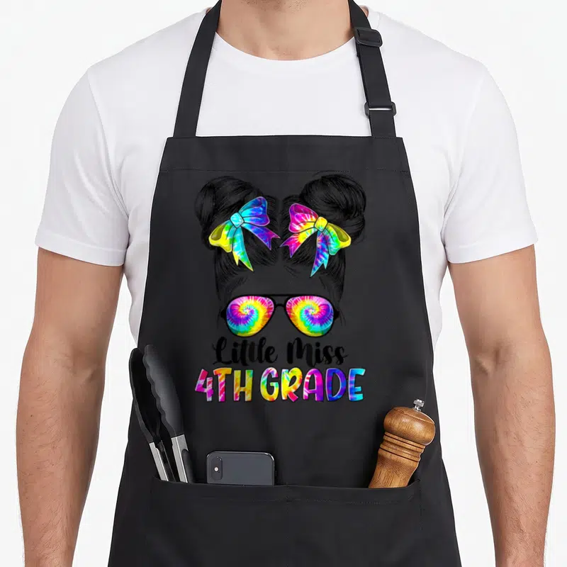 Miss 4th tie-dye apron - Cartoon girl with two buns, rainbow bows, sunglasses, and Little Miss 4TH GRADE text — shown on a black cotton canvas apron against a white background