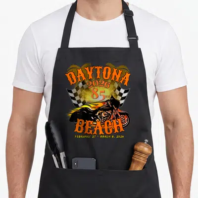 85th florida apron - Retro 2026 Daytona 85th motorcycle with flames, checkered flag, and palm trees in orange and yellow — shown on a black cotton canvas apron against a white background