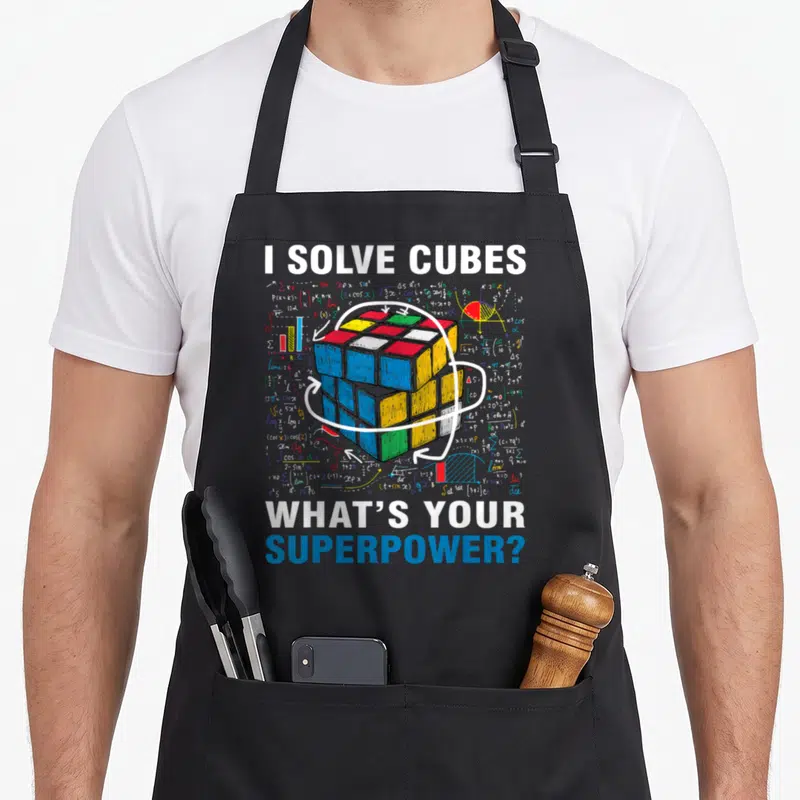 Solve rubiks cube apron - Cartoon Rubik's cube with math formulas, three rotation arrows, and text I SOLVE CUBES WHAT'S — shown on a black cotton canvas apron against a white background