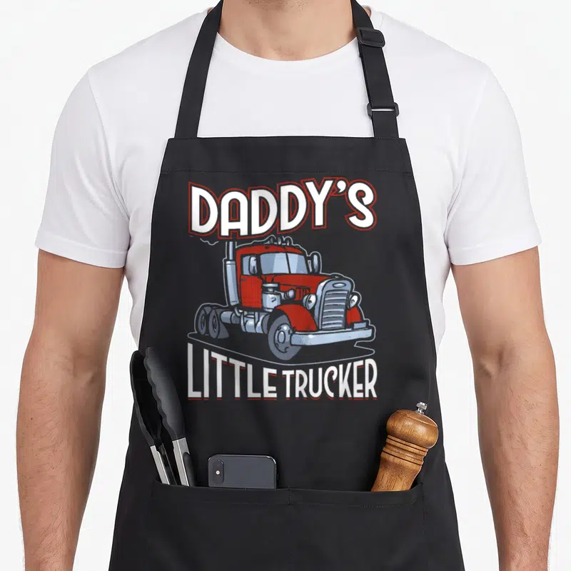 Daddy truck apron - Red cartoon semi-truck with arched DADDY'S LITTLE TRUCKER text in white and grey — shown on a black cotton canvas apron against a white background