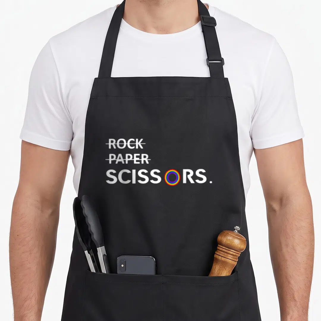 Lgbt paper apron - Rock Paper Scissors text with rainbow O and crossed out words — shown on a black cotton canvas apron against a white background