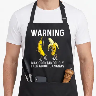Fruit banana apron - Two yellow cartoon bananas with white WARNING text and stacked slogan below — shown on a black cotton canvas apron against a white background