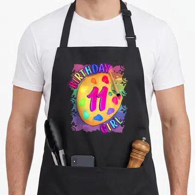 Painting 11th birthday apron - Yellow palette with pink 11, paintbrush, and BIRTHDAY GIRL text surrounded by colorful — shown on a black cotton canvas apron against a white background