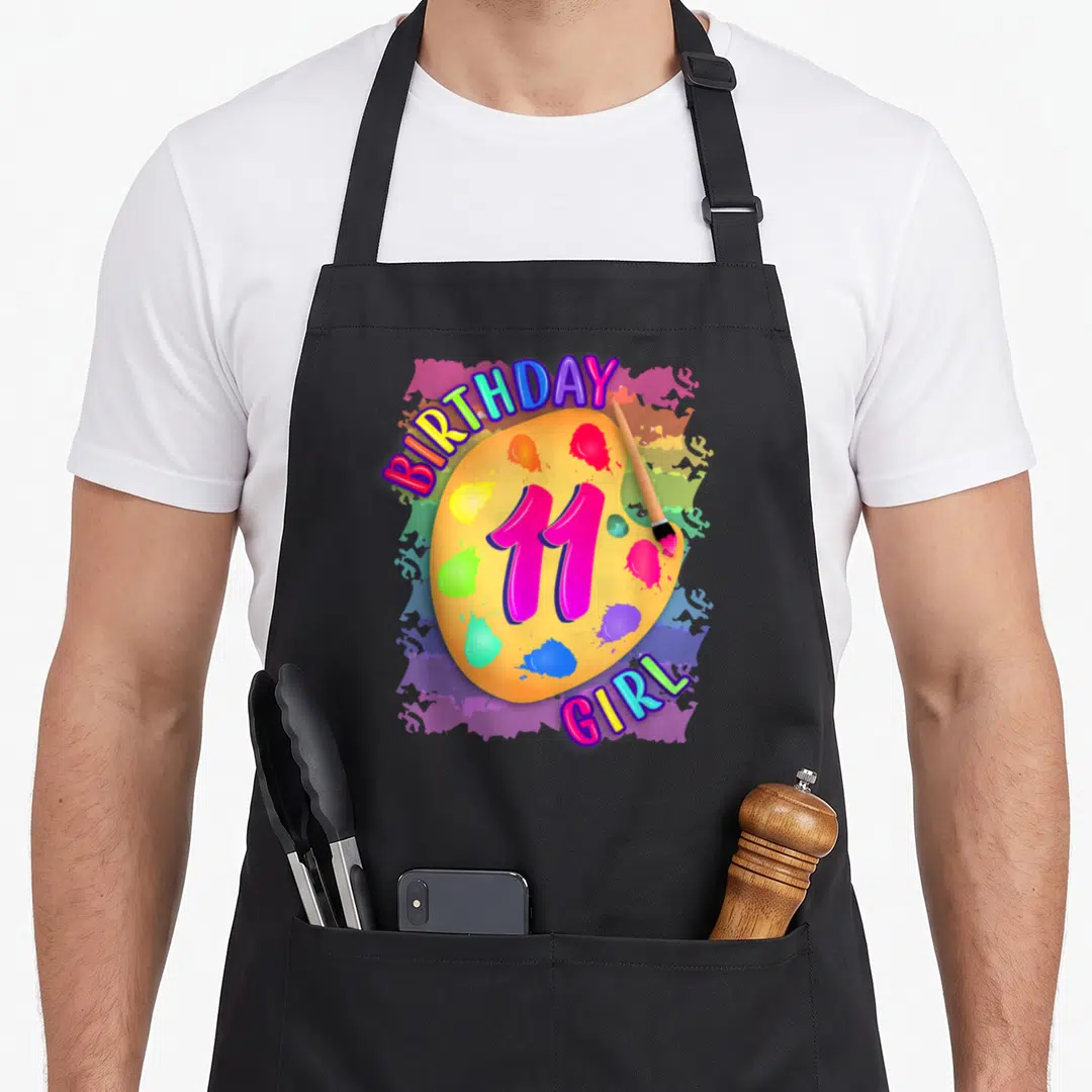 Painting 11th birthday apron - Yellow palette with pink 11, paintbrush, and BIRTHDAY GIRL text surrounded by colorful — shown on a black cotton canvas apron against a white background