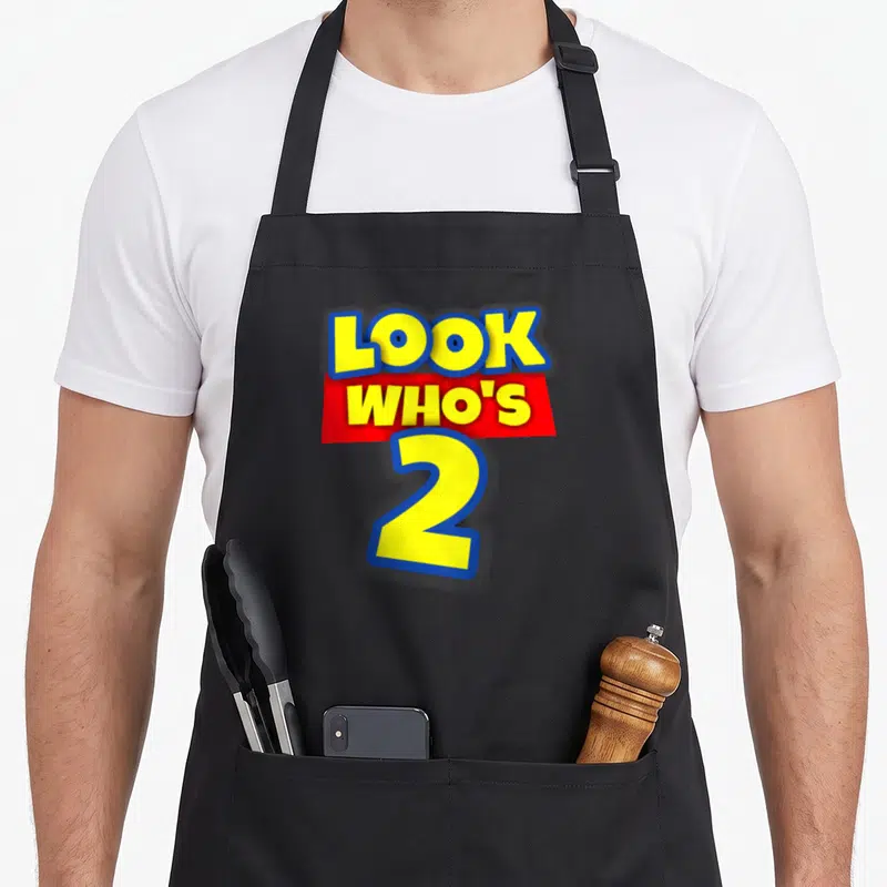 Funny theme birthday apron - Yellow text reading Look Who's 2 with blue outlines and red banner — shown on a black cotton canvas apron against a white background