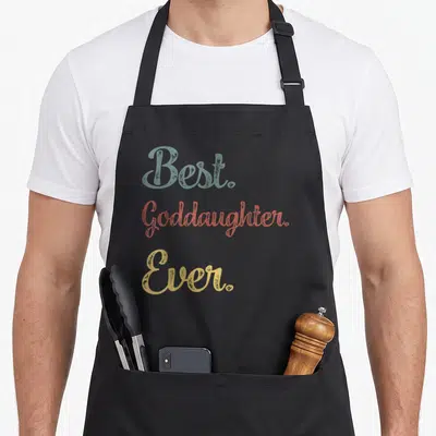 Funny goddaughter apron - Best. Goddaughter. Ever. text in blue-grey, coral, and gold stacked typography — shown on a black cotton canvas apron against a white background